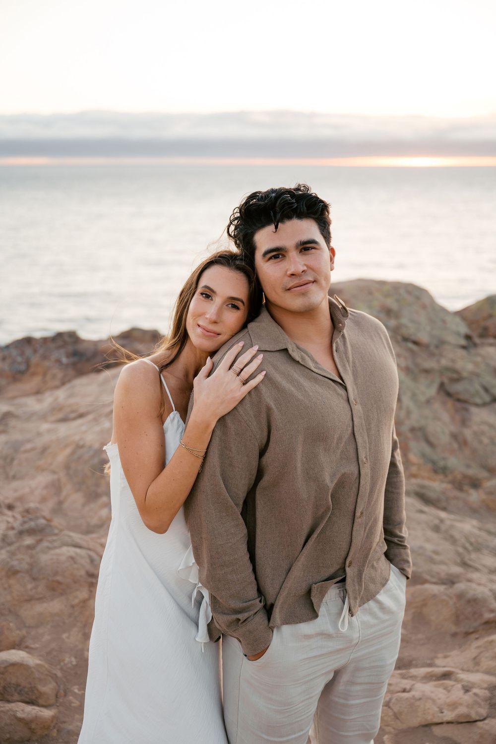A couple shares romantic moments at sunset on rocky coastal cliffs in warm earth tones.