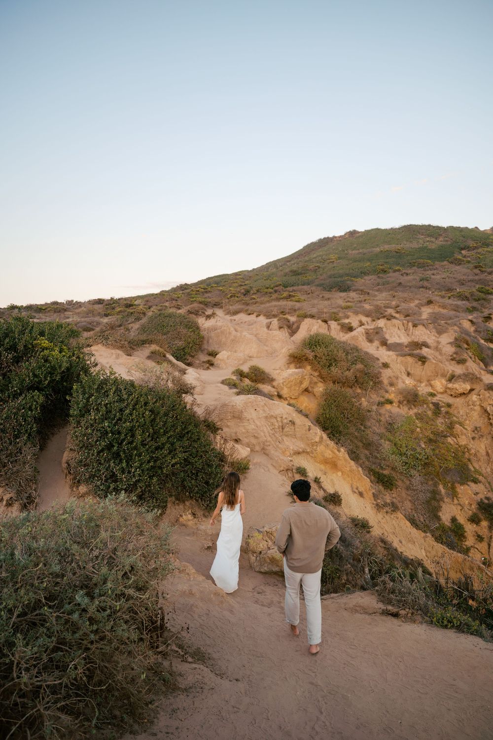 Two people walk along a winding dirt path on a coastal cliff during golden hour.