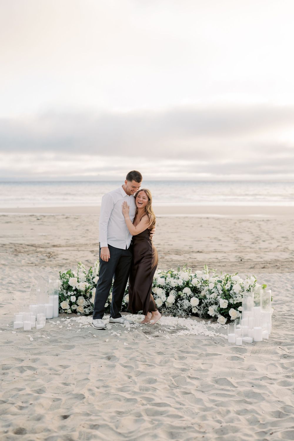 A couple shares a romantic moment surrounded by white floral arrangements and candles on a sandy beach at sunset.
