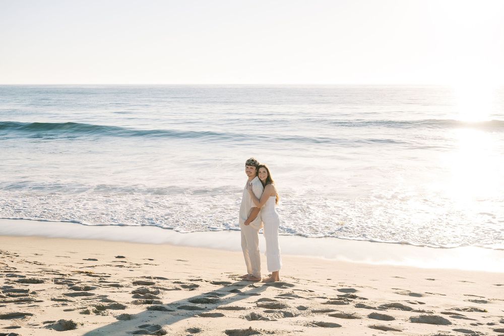 Couple walking along a serene beach at sunset with waves crashing on the shore and golden light illuminating the scene.