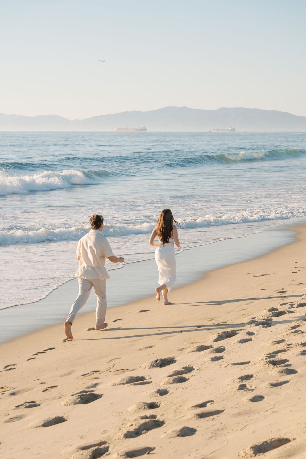 Two people in white outfits run playfully along a sandy beach with rolling waves and mountains in the background.