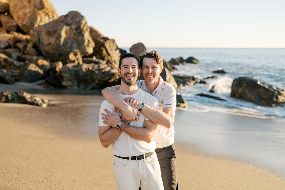 A loving couple embraces on a sandy beach with rocky cliffs and ocean waves in the background at sunset.