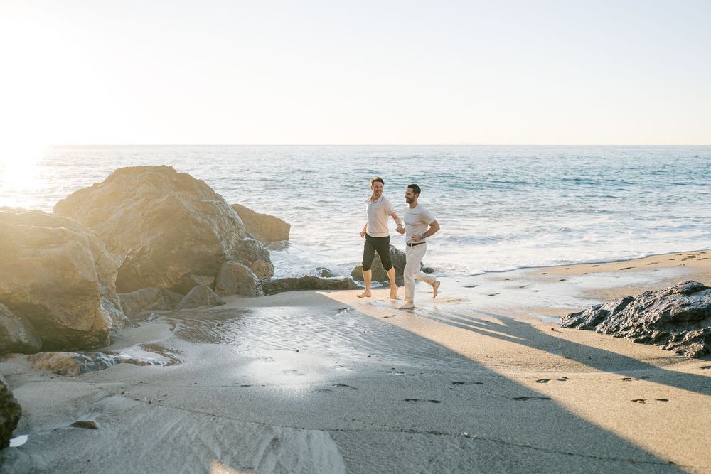 People enjoying a scenic beach walk along rocky coastline during golden hour with waves crashing against cliffs.