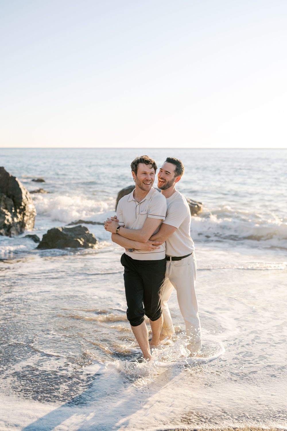 Two people joyfully playing and splashing in the ocean waves at sunset along a scenic beach.