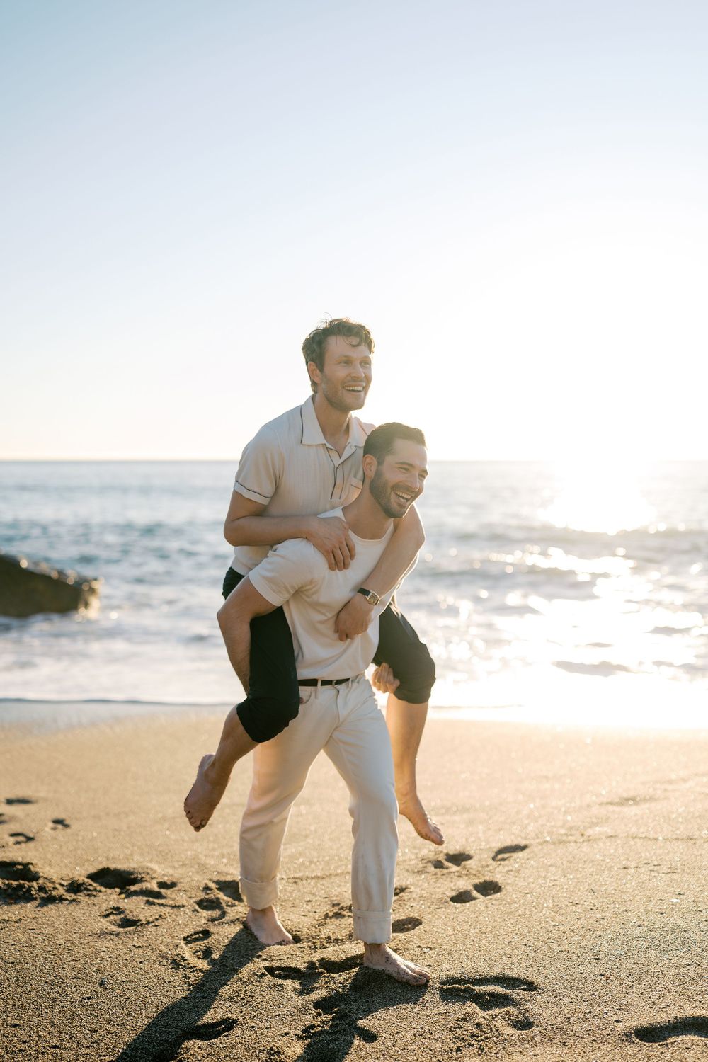 A couple shares a playful moment together on a sunny beach at sunset with golden light illuminating the sand.
