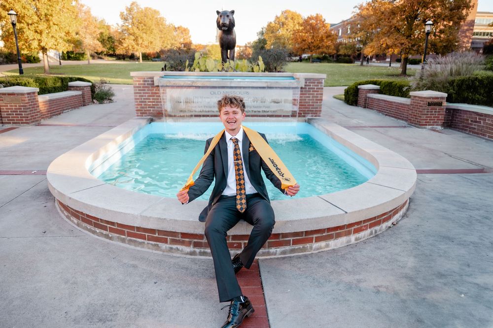 Graduate posing by a campus fountain in formal attire with academic regalia.