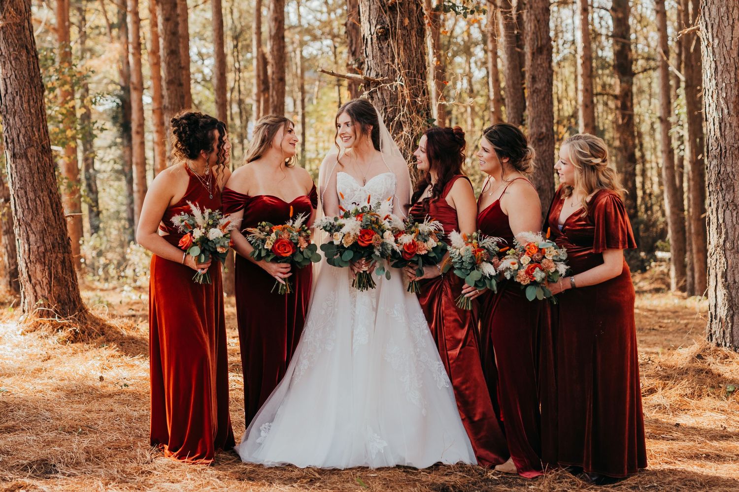 Wedding party in burgundy dresses poses together in a sunlit forest setting with pine trees in the background.