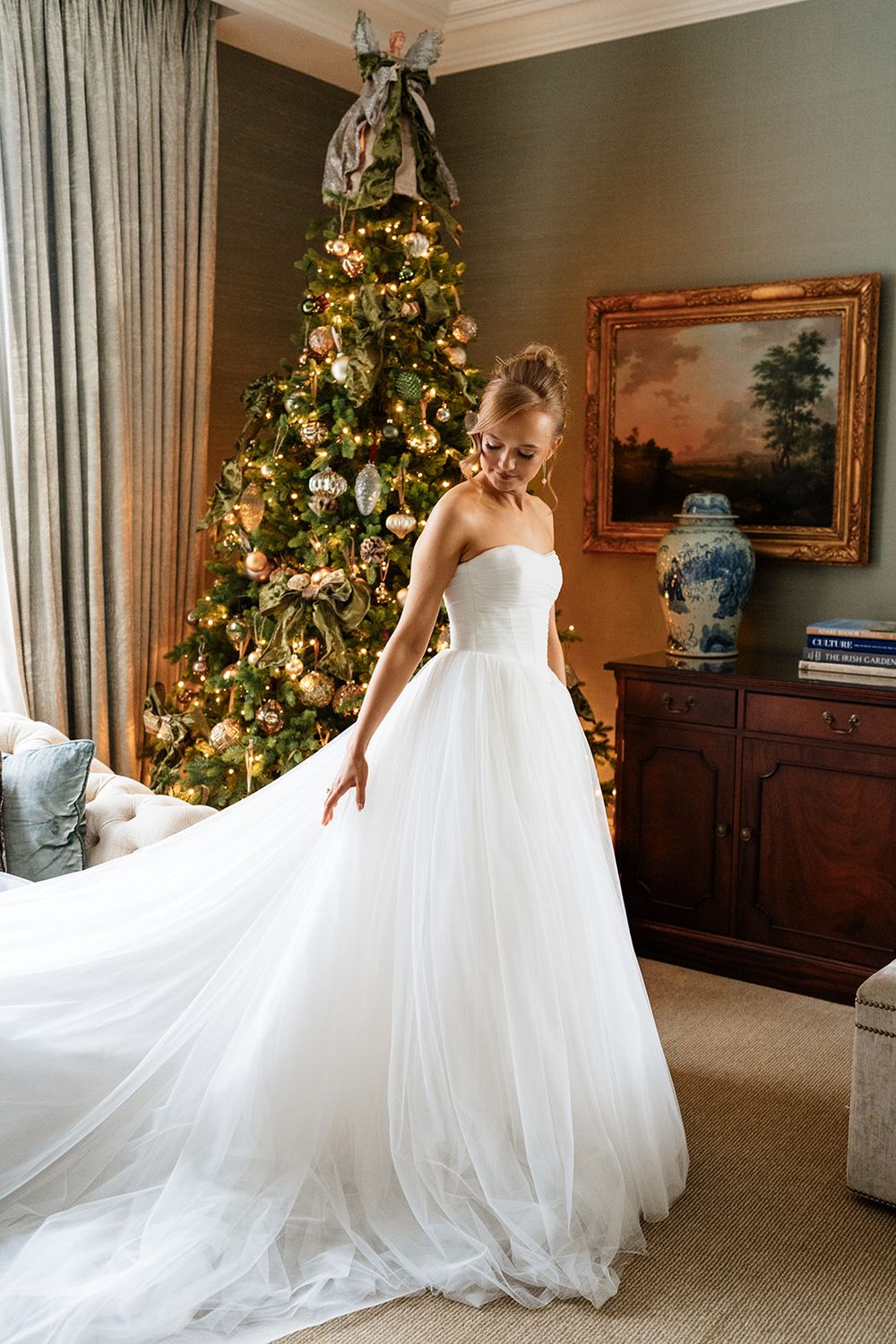 A bride in a flowing white wedding gown twirls elegantly in front of decorated Christmas trees in an elegant room.