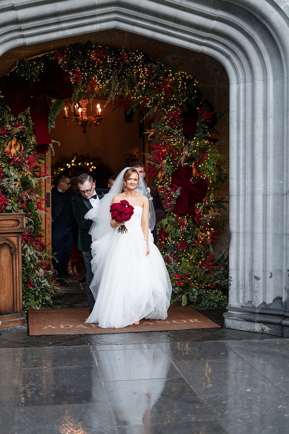 A bride in a flowing white gown twirls and dances under a stone archway decorated with red climbing roses.
