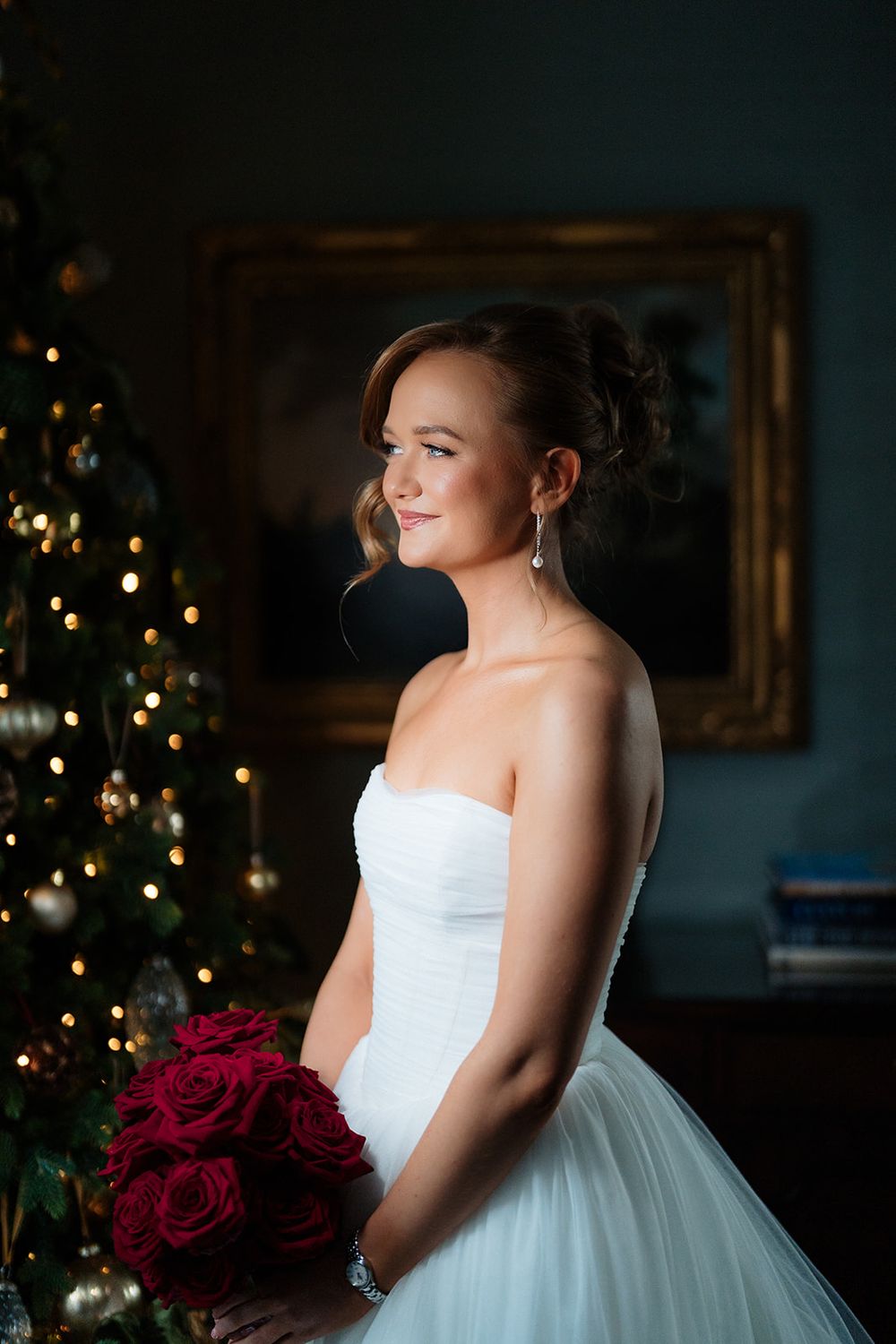 A bride in a white strapless gown holds red roses beside a Christmas tree in an elegant indoor setting.