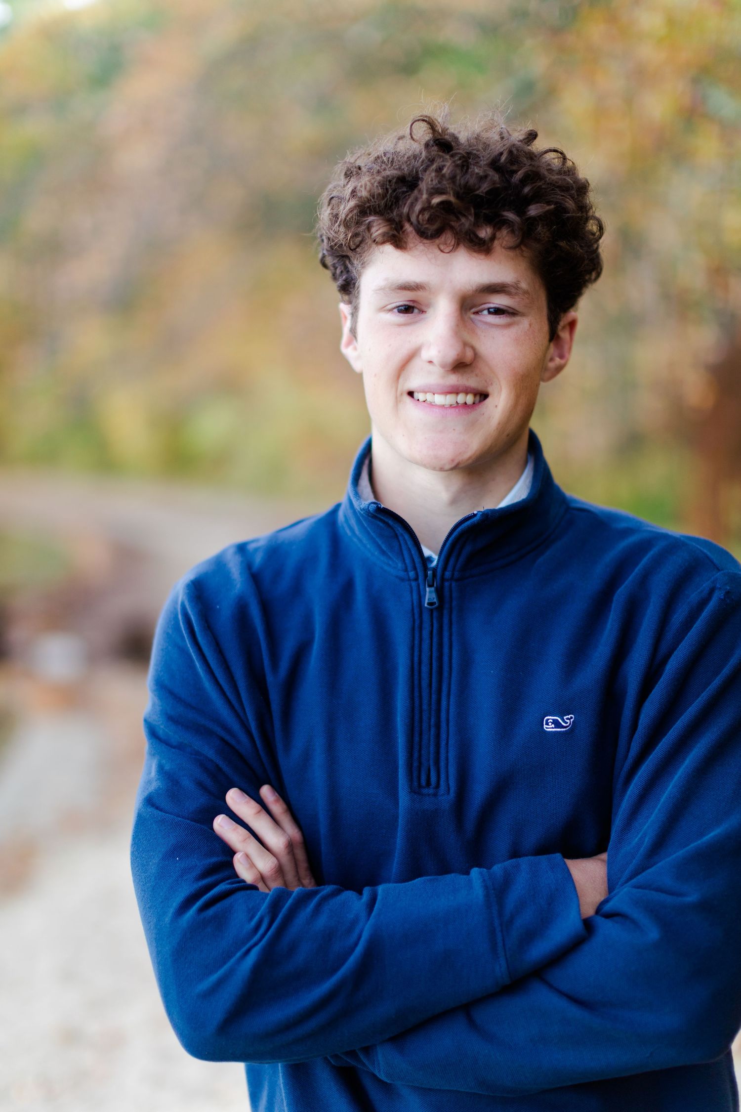 A person in a navy blue fleece jacket poses outdoors with crossed arms and curly hair against a blurred autumn background.