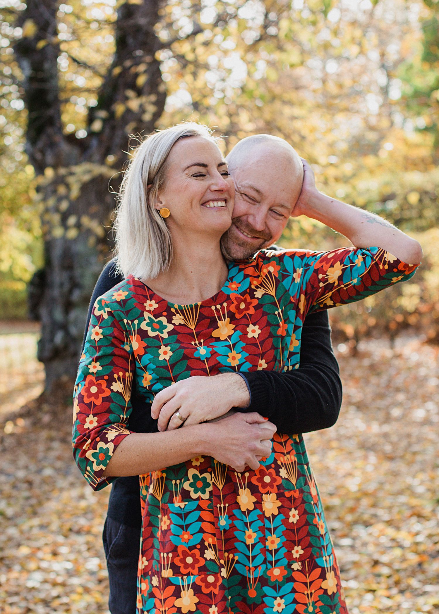 Happy couple hugging and laughing in autumn park, woman in colorful floral dress with fall foliage background.