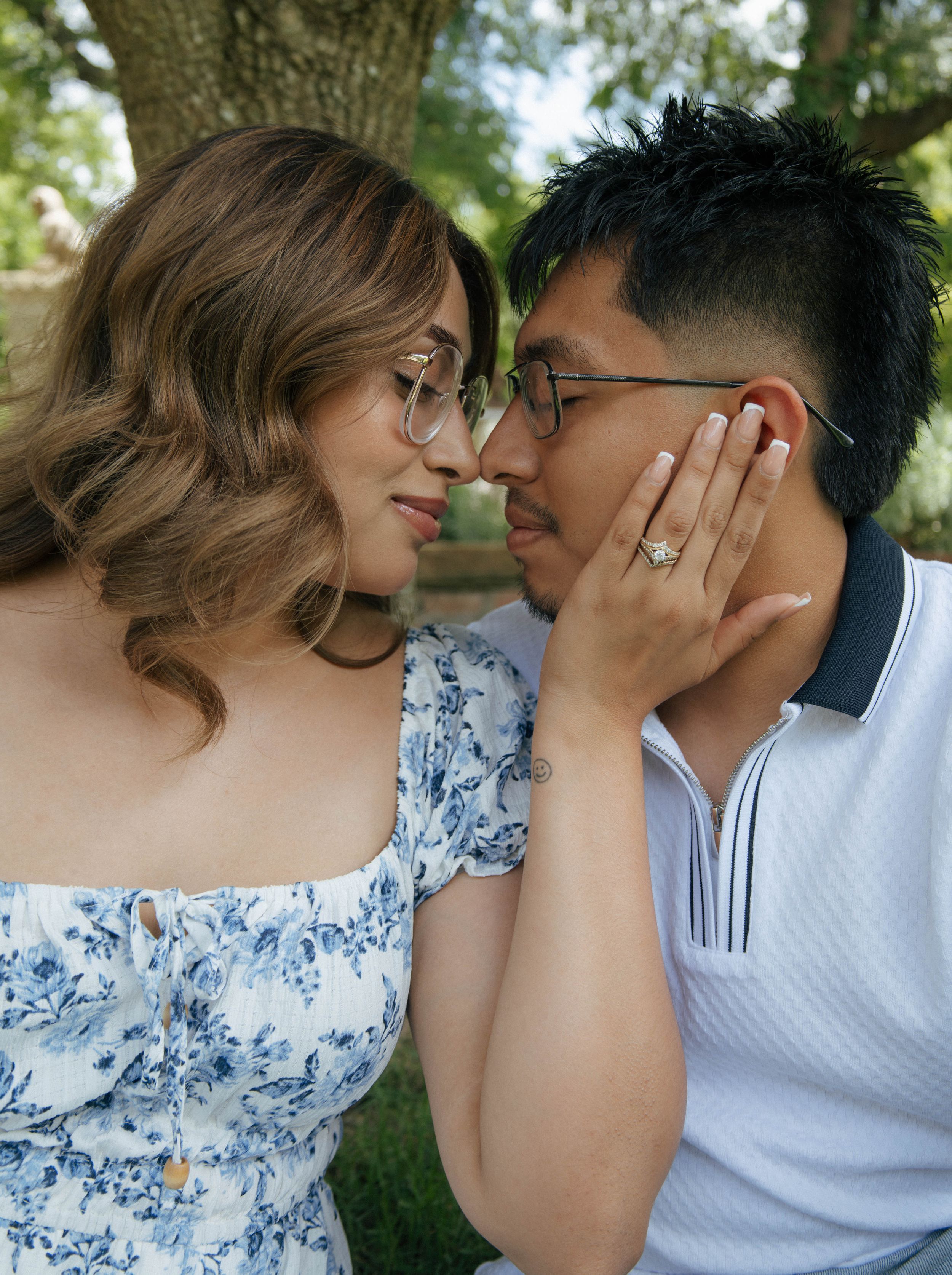 Couple in glasses sharing a romantic moment outdoors, woman wearing floral dress and engagement ring.