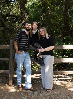 Family of three posing outdoors by a wooden fence, surrounded by lush green trees on a sunny day.