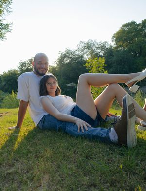Couple relaxing on green grass outdoors in golden hour sunlight, smiling at camera near trees.