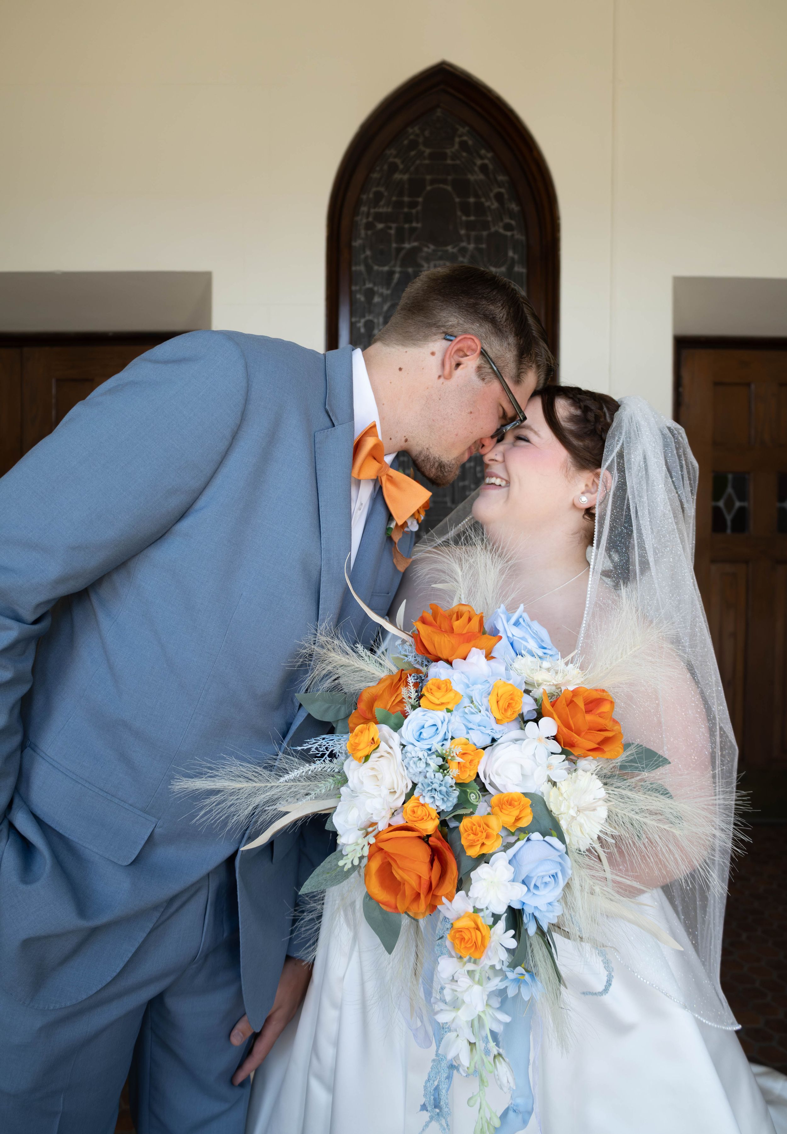 A bride and groom share a romantic moment in a church, with orange and blue floral bouquet and matching blue suit and orange bow tie.
