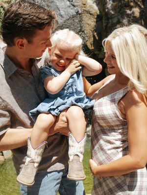 Happy family with father holding toddler girl in blue dress while mother smiles outdoors on sunny day.