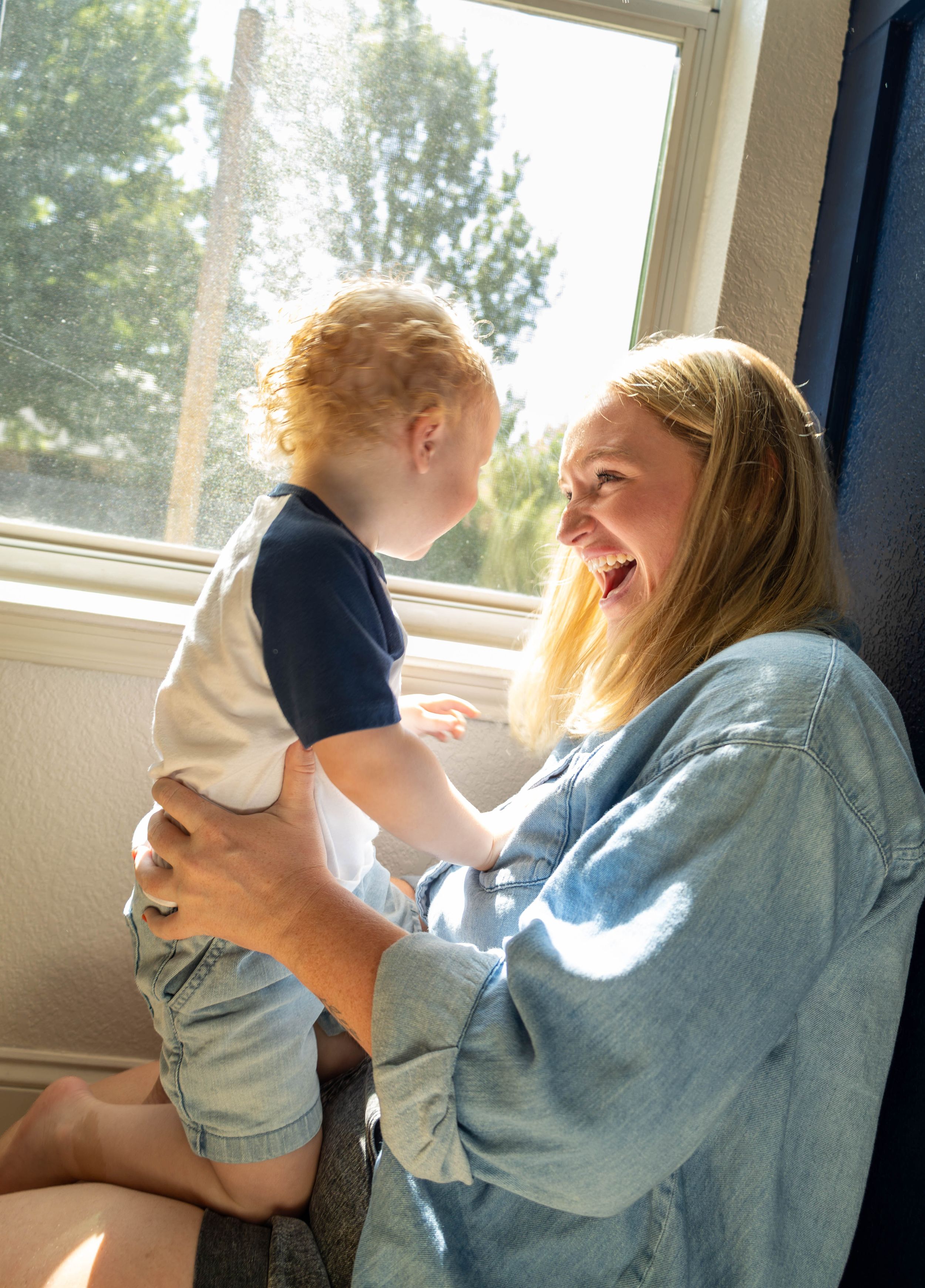 A joyful mother holds her red-haired toddler boy near a sunny window, both laughing and sharing a playful moment.