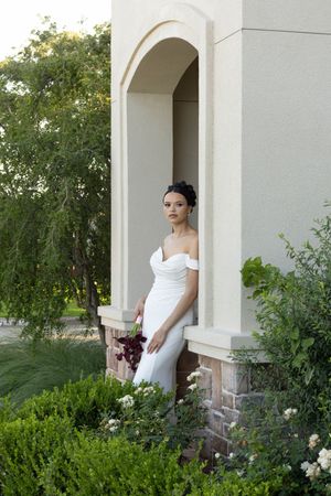 Bride in white off-shoulder gown holding dark bouquet, posed in arched alcove surrounded by lush greenery.