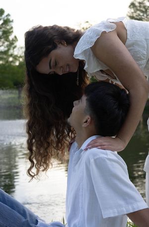 A couple in white clothing share a romantic moment by a serene lake, with the woman leaning toward the man.
