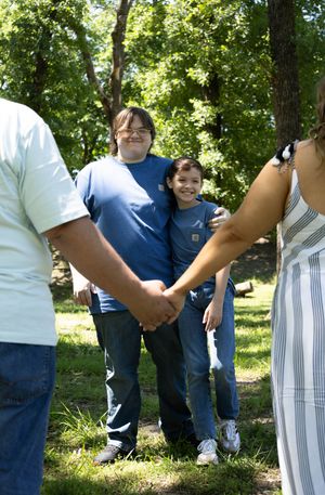 Two boys in blue shirts smile outdoors in a park, surrounded by family holding hands on a sunny day.