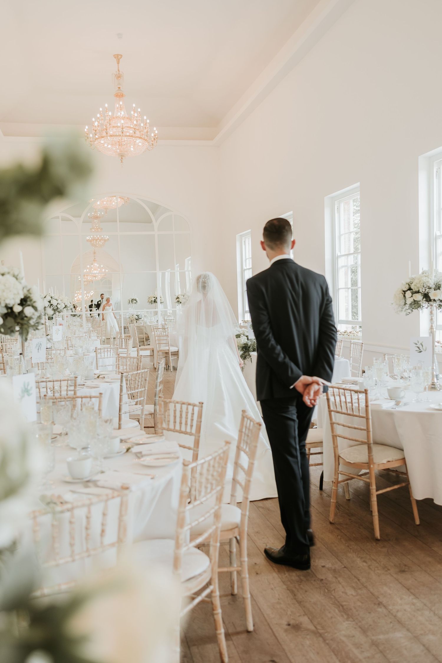 Elegant wedding reception venue with white chairs, crystal chandelier and floral arrangements in a bright room.