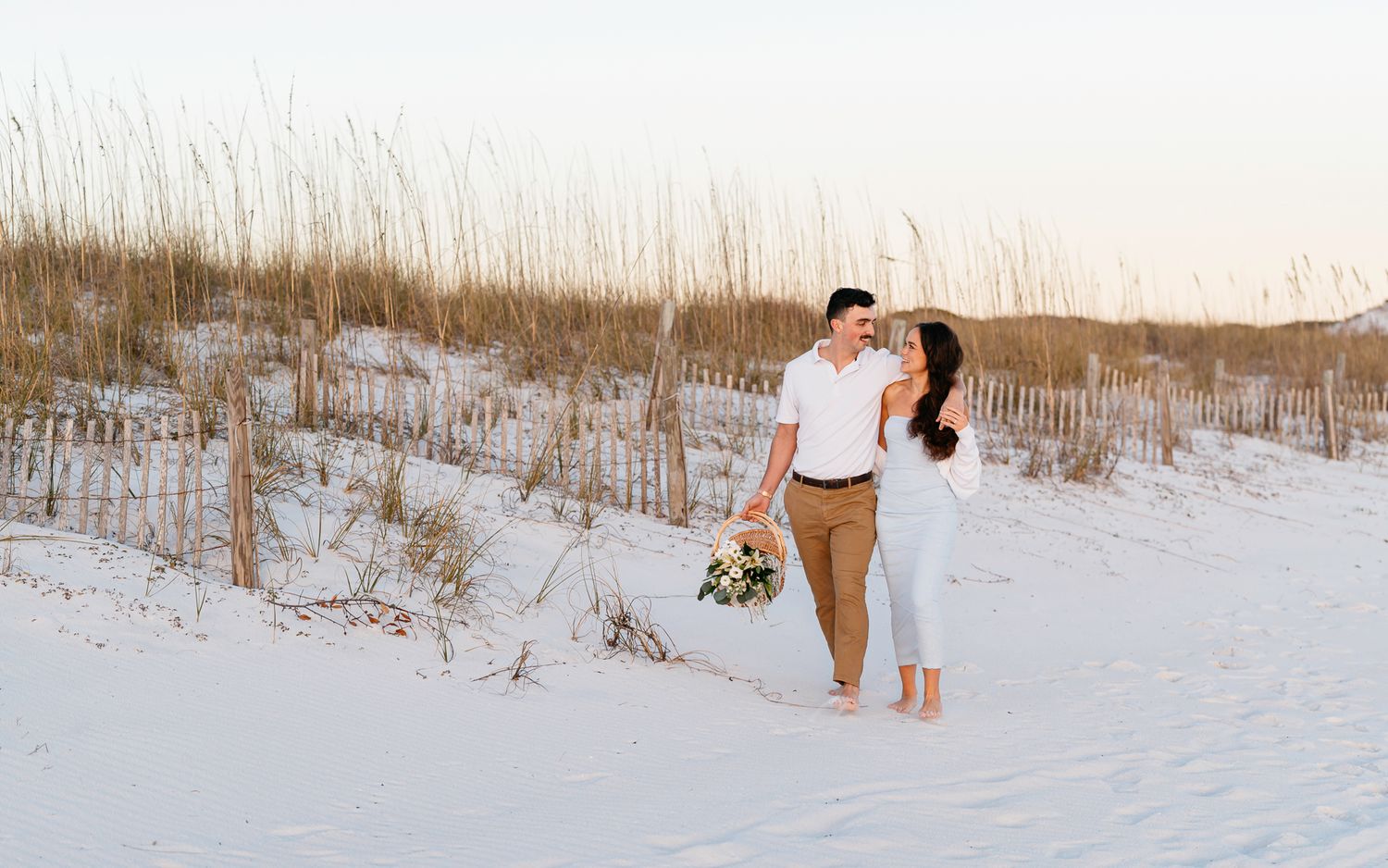 A couple shares a romantic moment on white sand dunes at sunset with beach grass in the background.