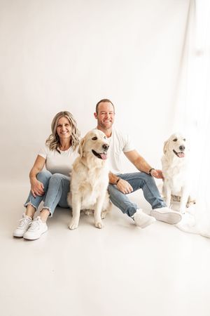 A couple sits on a white floor with three golden retriever dogs in a minimalist photo studio setting.