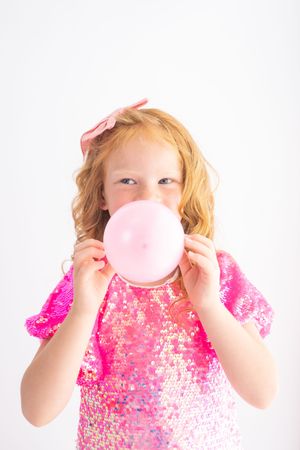 A young girl in a pink sequin dress and bow blows up a pink balloon against a white background.