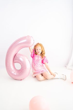 A smiling young girl in a pink sequin dress sits beside a large pink number 6 balloon on a white background.