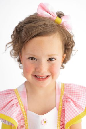 Smiling young girl wearing a pink gingham dress with yellow trim and a pink bow in her curly brown hair.