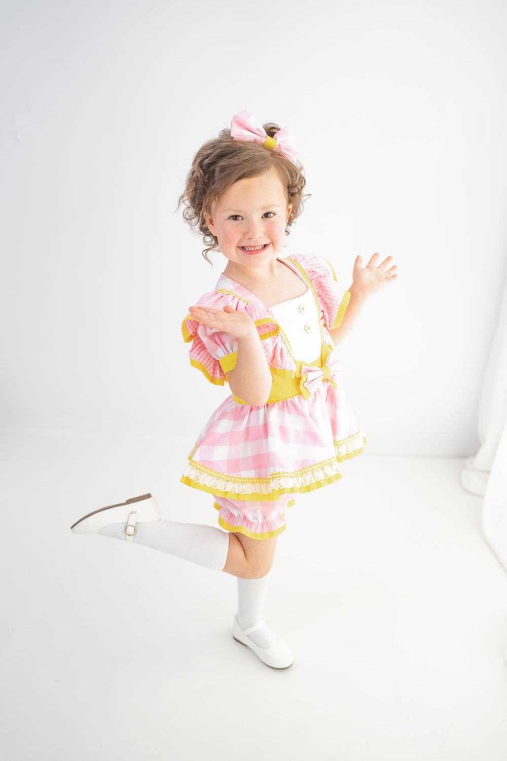 A cheerful toddler girl in a pink and yellow gingham dress with white boots poses playfully in a bright studio.
