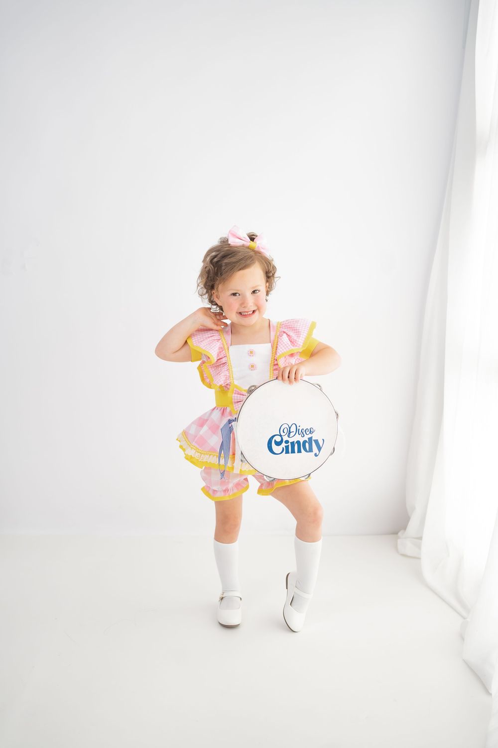 Young girl in pink ruffled dress holding a drum labeled Disco Cindy, wearing white boots, smiling in white studio.
