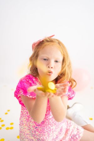 Young redhead girl in pink sequin dress blowing gold confetti from her hands at a birthday party.