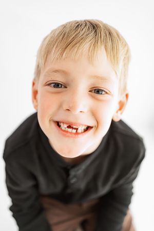 Smiling blonde boy with missing front teeth wearing a dark shirt against a white background.