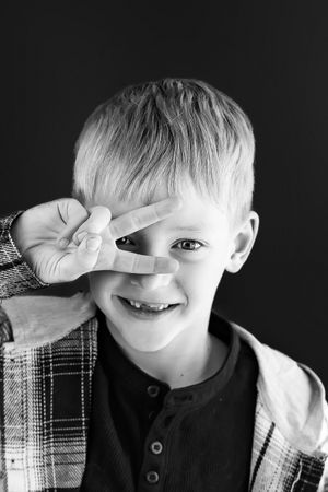 Black and white portrait of a smiling young boy making a peace sign over his eye.