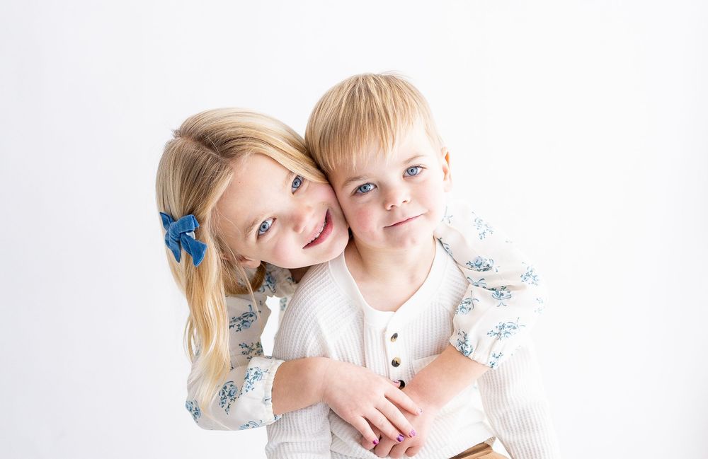 Three sweet siblings in white clothing share affectionate moments together against a light background.