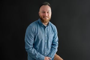 Professional headshot of someone wearing a light blue button-down shirt against a dark background.