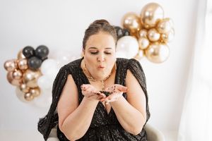 Woman in black sequin dress blowing glitter with gold, black, and rose gold balloons in background.
