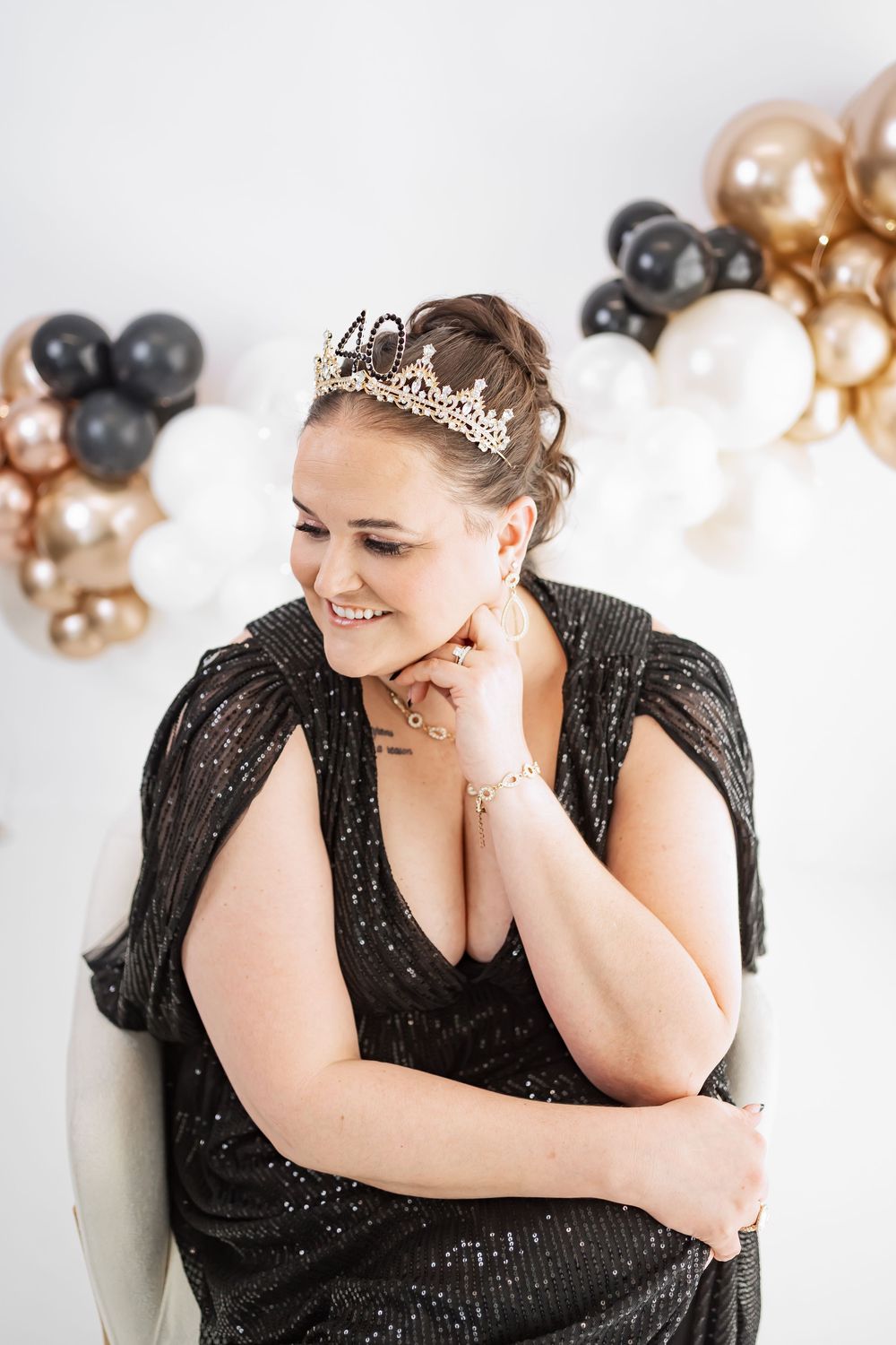 Woman in black sequin dress and gold tiara smiles at 40th birthday party with black and gold balloons.