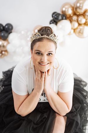 Woman wearing a 40th birthday tiara crown smiles in black tulle skirt with gold and black balloon backdrop.