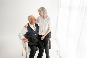 Two people in white and black attire share a lighthearted moment while sitting together in a bright studio setting.