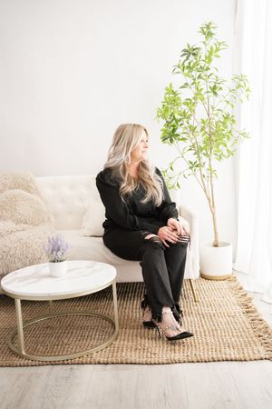 A person in black clothing sits on a jute rug next to a white round table with indoor plants in a bright room.