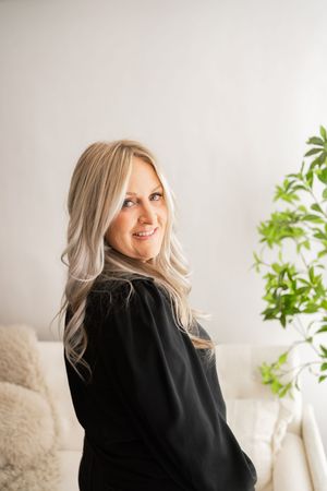 Woman with platinum blonde hair wearing black sweater smiles while looking up in bright minimalist setting with plants.