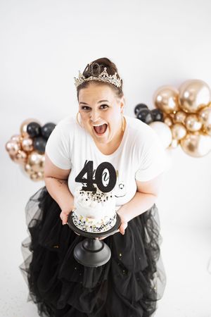 Woman celebrating 40th birthday holding cake, wearing tiara and black tulle skirt, with gold and black balloons.