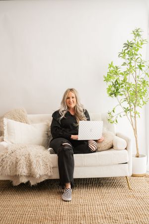 Cozy home office setup with white couch, furry throw blanket, potted plant and laptop against neutral wall.