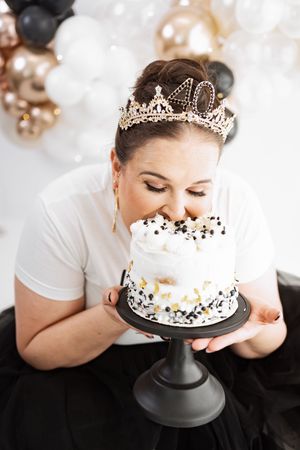 Woman wearing a 40th birthday tiara biting into a white cake with black and gold decorations at a balloon party.