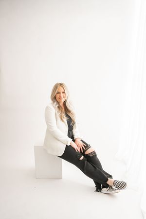 Professional studio portrait of someone wearing a white blazer and black pants sitting against white background.
