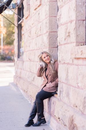 A person in a brown sweater and black pants sits casually against a textured stone wall outdoors.