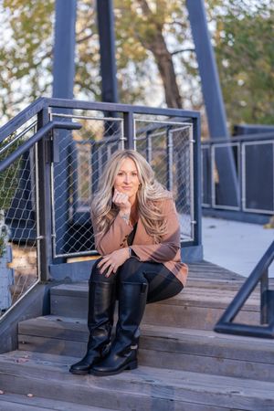 A person in black boots and casual wear poses playfully on wooden stairs with metal railings outdoors.